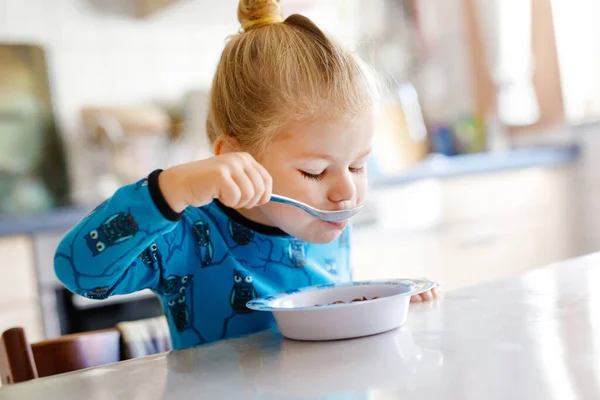 Linda menina criança comendo cereais saudáveis com leite para o café da manhã. Bonito bebê feliz criança em roupas coloridas sentado na cozinha e se divertindo com a preparação de aveia, cereais. Interior em casa — Fotografia de Stock