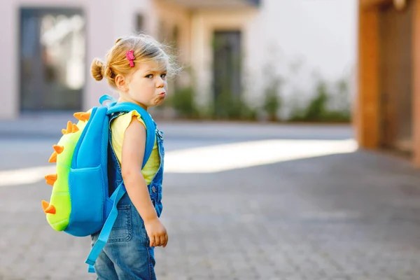 Linda niñita adorable en su primer día yendo a la escuela de juegos. Saludable bebé triste molesto caminando a la guardería. Miedo al jardín de infantes. Niño infeliz con mochila en la calle de la ciudad, al aire libre —  Fotos de Stock