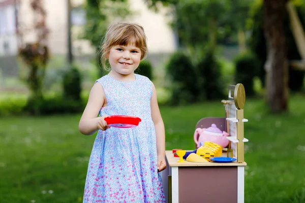 Little preschool girl playing with toy kitchen in garden. Happy toddler child having fun with role activity game preparing food. Children play outdoors in summer. — Stock Photo, Image