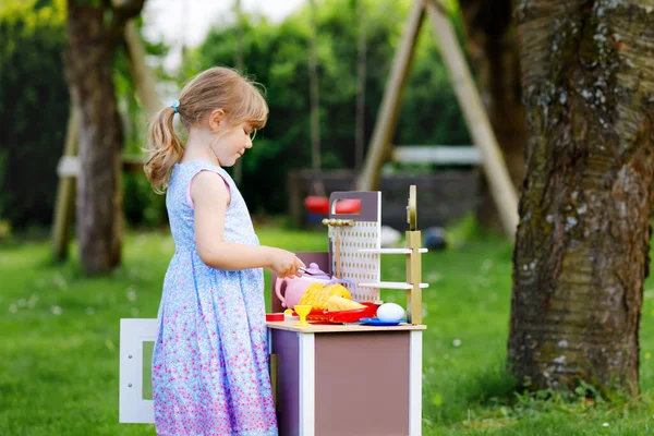 Pequena menina pré-escolar brincando com cozinha de brinquedo no jardim. Criança feliz se divertindo com o jogo de atividade de papel preparando alimentos. Crianças brincam ao ar livre no verão. — Fotografia de Stock