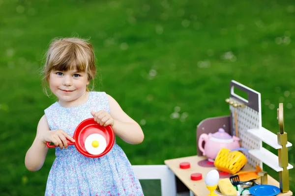 En liten förskoletjej som leker med leksaksköket i trädgården. Glad småbarn har kul med roll aktivitetsspel förbereda mat. Barn leker utomhus på sommaren. — Stockfoto