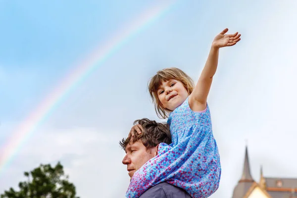 Маленькая дошкольница сидит на плече отца. Счастливый малыш и мужчина наблюдают радугу на небе после летнего дождя. Счастливая семья, дружба, любовь. Летом. Папа и дочь. День отца. — стоковое фото