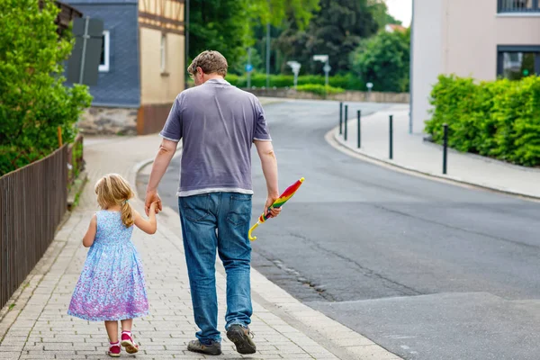 小さな就学前の女の子と父親が街を歩いています。幸せな幼児の子供と男が手を握っている。幸せな家族、絆、愛。夏だ。パパと娘よ。父の日だ。後ろから. — ストック写真