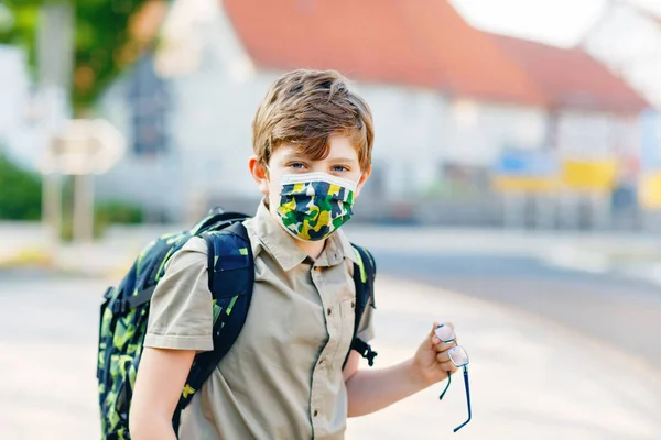 Glücklicher Junge mit Brille und medizinischer Maske wegen Coronavirus-Pandemie. Schulkind mit Schulranzen wartet am sonnigen Tag auf dem Schulweg auf den Bus. Gesundes Kind auf der Straße. — Stockfoto