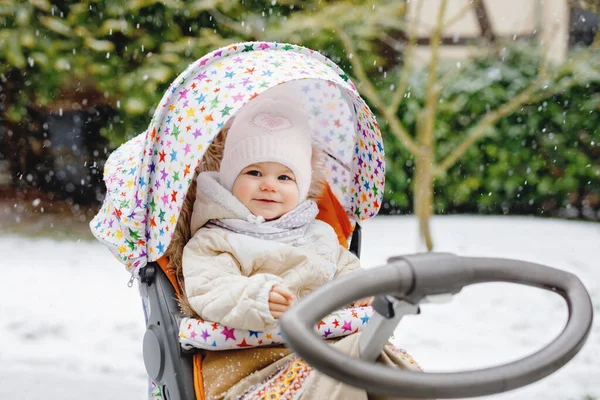 Jolie petite belle petite fille assise dans la poussette ou la poussette par une froide journée d'hiver enneigée. Joyeux enfant souriant dans des vêtements chauds, mode manteau de bébé élégant. Babys première neige. Promenade hivernale en plein air. — Photo