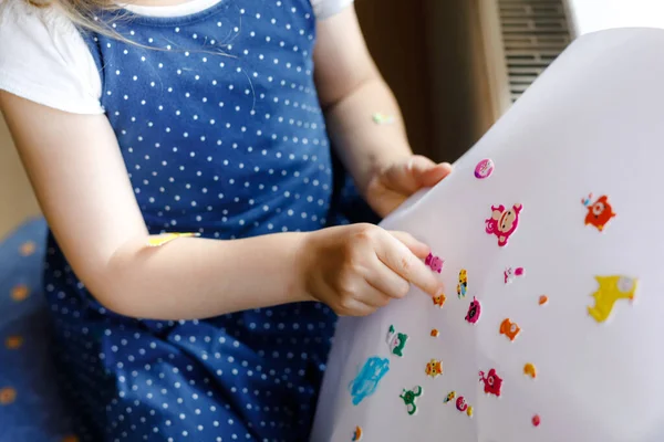 Liten småbarn flicka leker med olika färgglada djur klistermärken. Begreppet aktivitet hos barn under karantän för koronavirus. Glad rolig barn att ha kul med stick klistermärken i ansiktet. — Stockfoto