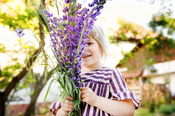 Nettes entzückendes kleines Mädchen mit riesigem Strauß blühender violetter Lupniesblüten. Porträt eines lächelnden Vorschulkindes im heimischen Garten an warmen Frühlingstagen oder Sommertagen. Sommerzeit. — Stockfoto