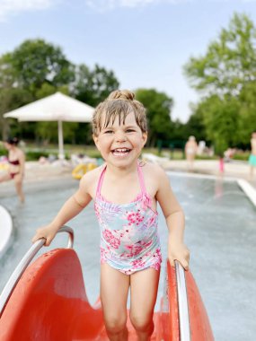Küçük bir anaokulu kızı ılık bir yaz gününde açık havuzda su sıçratıyor. Mutlu sağlıklı çocuk şehir havuzunda güneşli havanın tadını çıkarıyor. Suyla dışarıda çocuk aktivitesi.