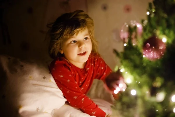 Piccola ragazza bambino carino a letto sotto l'albero di Natale e sognando di Babbo Natale a casa, all'interno. Festa cristiana tradizionale. Bambino felice in attesa di regali a Natale. Accogliente luce morbida — Foto Stock