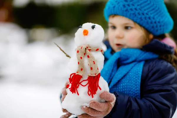 Χαριτωμένο κοριτσάκι που φτιάχνει μίνι χιονάνθρωπο και τρώει μύτη καρότου. Αξιολάτρευτο υγιές χαρούμενο παιδί που παίζει και διασκεδάζει με το χιόνι, σε εξωτερικούς χώρους την κρύα μέρα. Ενεργός ελεύθερος χρόνος με τα παιδιά το χειμώνα — Φωτογραφία Αρχείου