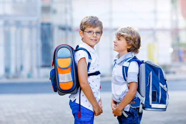 Two little kid boys with backpack or satchel. Schoolkids on the way to school. Healthy adorable children, brothers and best friends outdoors on the street leaving home. Back to school. Happy siblings.