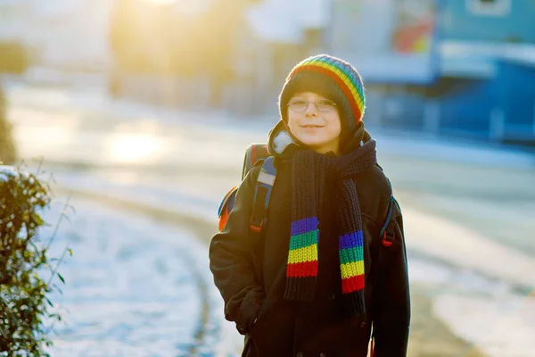 Chłopiec z podstawówki, idący do szkoły podczas opadów śniegu. Szczęśliwe zdrowe dziecko w okularach zabawy i zabawy ze śniegiem. Z plecakiem lub torbą w kolorowe zimowe ubrania. — Zdjęcie stockowe