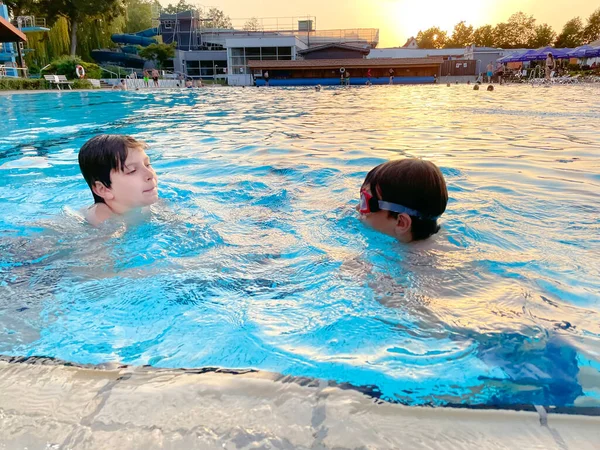 暖かい夏の日に屋外スイミングプールで遊んでいる2人の学校の子供たち。市の公共プールで天気の良い日を楽しむ幸せな健康的な子供たち。水で屋外の子供の活動. — ストック写真