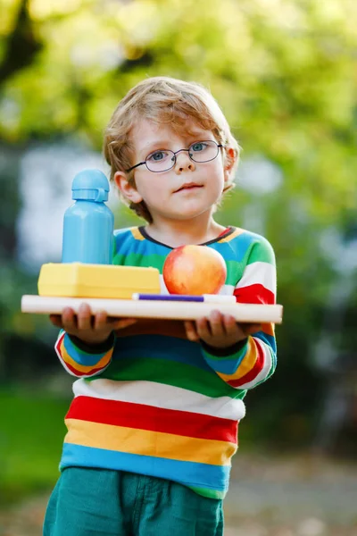 Glad liten förskolekille med böcker, äpple och dricka flaska på sin första dag till grundskolan eller förskolan. Leende barn, student med glasögon, utomhus. Tillbaka till skolans utbildningsidé. — Stockfoto