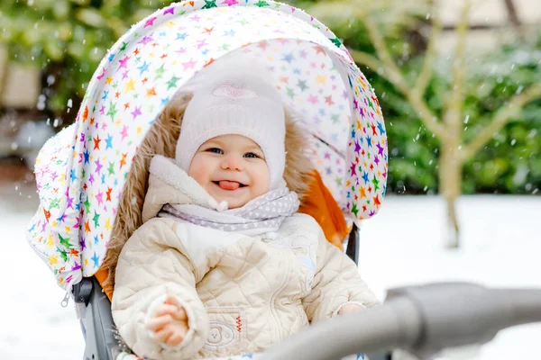 Cute little beautiful baby girl sitting in the pram or stroller on cold snowy winter day. Happy smiling child in warm clothes, fashion stylish baby coat. Babys first snow. Winter walk outdoors. — Stock Photo, Image