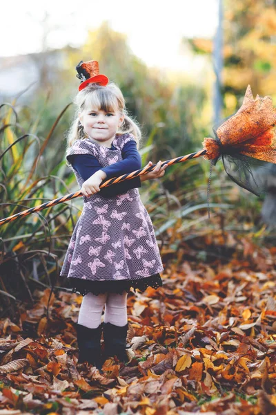 Linda niña pequeña vestida como una bruja celebra Halloween. Niño feliz al aire libre, con sombrero naranja divertido y escoba de bruja. Hermosa temporada de festivales familiares en octubre. Actividades al aire libre —  Fotos de Stock