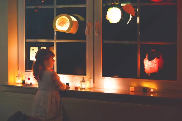Menina criança sentada à janela com lanternas artesanais feitas à mão com velas para procissão de St. Martin. Criança olhando para lanterna brilhante. Tradição alemã Martinsumzug. Decoração de casa — Fotografia de Stock