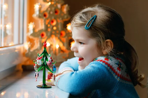 Niña pequeña sentada junto a la ventana y decorando un pequeño árbol de Navidad de vidrio con pequeños juguetes de Navidad. Feliz niño sano celebrar la fiesta tradicional familiar. Adorable bebé.. — Foto de Stock