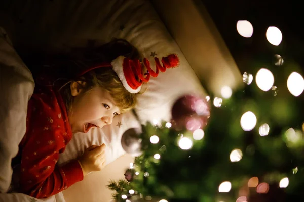 Pequena menina bonito na cama sob a árvore de Natal e sonhando com o Papai Noel em casa, dentro de casa. Festival cristão tradicional. Criança feliz esperando presentes em xmas. Luz suave aconchegante — Fotografia de Stock