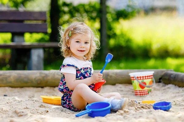 Schattig peuter meisje spelen in zand op buiten speeltuin. Mooie baby in rode broek die zich vermaakt op zonnige warme zomerdag. Kind met kleurrijk zandspeelgoed. Gezonde actieve baby buiten speelt spelletjes — Stockfoto