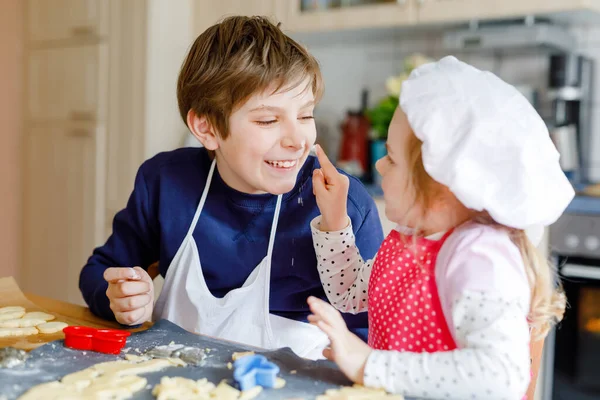 Linda niña pequeña y preadolescente niño hornear galletas de Navidad en casa en interiores. Niños, hermanos con delantal y sombrero de chef cortan galletas en la cocina doméstica. Hermano y hermana enamorados —  Fotos de Stock