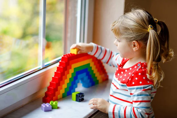 Cute little toddler girl by window create rainbow with colorful plastic blocks. People with rainbows around the world as protest and sign. — Stock Photo, Image