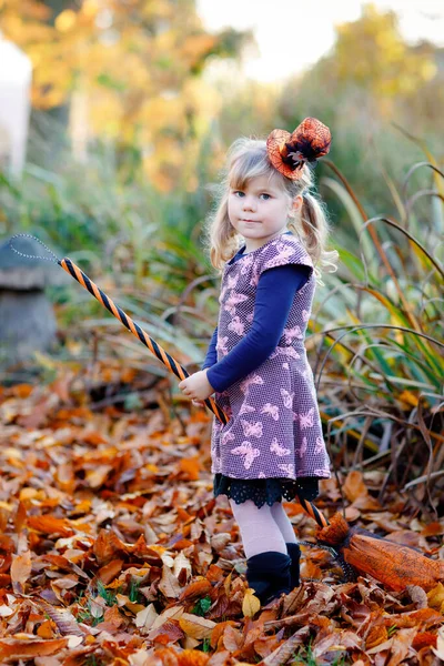 Linda niña pequeña vestida como una bruja celebra Halloween. Niño feliz al aire libre, con sombrero naranja divertido y escoba de bruja. Hermosa temporada de festivales familiares en octubre. Actividades al aire libre —  Fotos de Stock