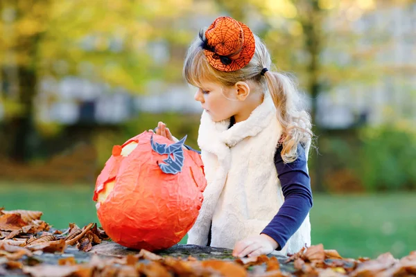 Cadılar Bayramı 'nda cadı kılığına girmiş küçük bir kız. Mutlu çocuk dışarıda, turuncu komik şapkası ve tatlı lanetli balkabağı çantasıyla. Ekim 'de aile festivali sezonu. Açık hava aktivitesi — Stok fotoğraf