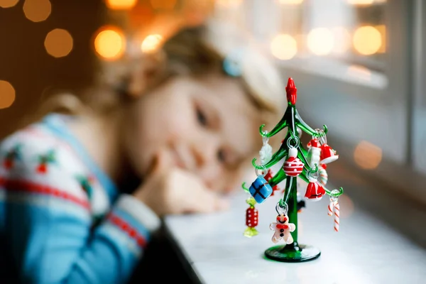 Niña pequeña sentada junto a la ventana y decorando un pequeño árbol de Navidad de vidrio con pequeños juguetes de Navidad. Feliz niño sano celebrar la fiesta tradicional familiar. Adorable bebé. Enfoque selectivo en el árbol — Foto de Stock