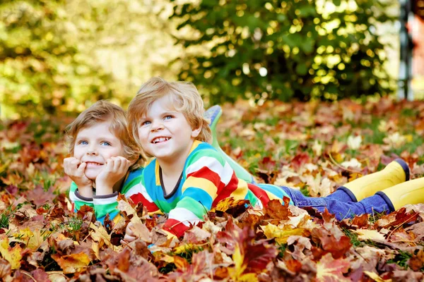 Two little twin kids boys lying in autumn leaves in colorful fashion clothing. Happy siblings having fun in autumn park on warm day. Healthy children with blond hairs and blue eyes with maple foliage. — Stock Photo, Image