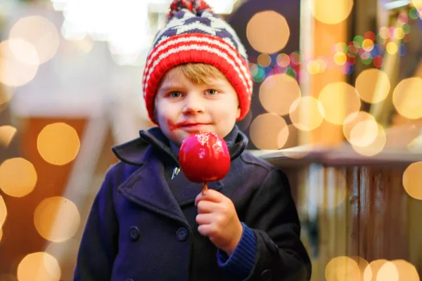 Mignon petit garçon mangeant de la pomme rouge sucrée sur le marché de Noël allemand. Joyeux enfant en vêtements d'hiver avec des lumières sur le fond. Un gamin qui regarde la caméra. Famille, tradition, concept de vacances. — Photo