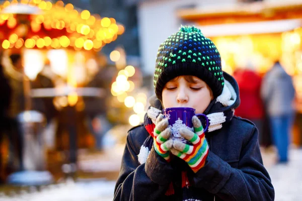 Niño lindo bebiendo ponche de niños calientes o chocolate en el mercado de Navidad alemán. Niño feliz en el mercado familiar tradicional en Alemania, niño risueño en ropa de invierno colorida — Foto de Stock