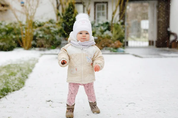 Счастливая маленькая девочка делает первые шаги на улице зимой через снег. Милый малыш учится ходить. Ребенок веселится в холодный снежный день. Первый снег, активность. Зимняя прогулка на природе — стоковое фото