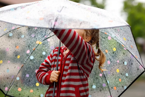 雨の日に大きな傘で遊んでいる小さな幼児の女の子。雨の中を走る幸せな正の子、水たまり。雨の服やゴムブーツを持つ就学前の子供。悪天候の日の子供の活動. — ストック写真