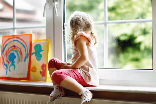 Schattig klein peutermeisje door raam met regenboog beschilderd met kleurrijke kleuren. Kleuterschool op slecht weer regenachtige dag kijkend uit het raam. Alleen overstuur kind — Stockfoto