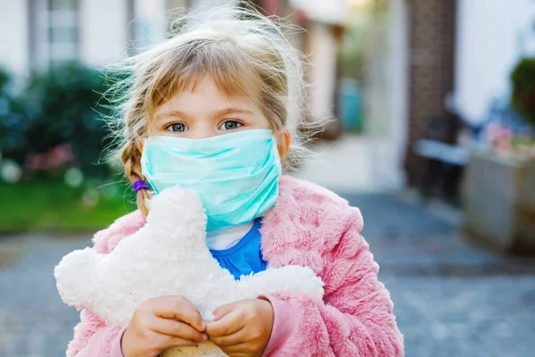 Menina pequena em máscara médica como proteção contra a doença de quarentena do coronavírus pandêmico. Criança bonito usando equipamentos de proteção como luta contra covid 19 e segurando urso brinquedo. — Fotografia de Stock