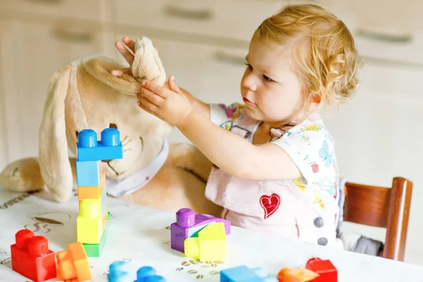 Adorable toddler girl with favorite plush bunny playing with educational toys in nursery. Happy healthy child having fun with colorful different plastic blocks at home. Cute baby learning creating. — Stock Photo, Image