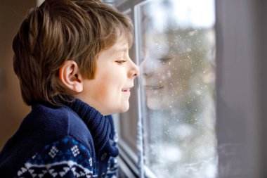 Mutlu sevimli çocuk pencerenin yanında oturuyor ve Noel sabahı ya da sabahleyin dışarıda karları seyrediyor. Gülümseyen çocuk kar yağışı ve büyük kar taneleriyle büyülenmiş.