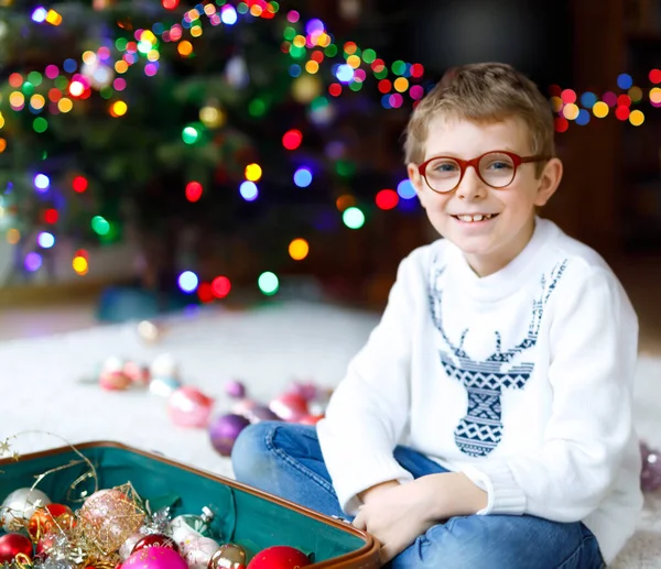 Schöner kleiner Junge mit Brille und buntem Vintage-Weihnachtsspielzeug und Bällen im alten Koffer. Kleines Kind, Schuljunge in festlicher Kleidung, die den Weihnachtsbaum schmückt. Kind lächelt in die Kamera. — Stockfoto