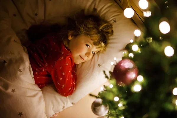 크리스마스 트리 아래 침대에 누워서 집안의 산타를 꿈꾸는 작고 귀여운 소녀. 전통 기독교 축제. 크리스마스 선물기다리는 행복 한아이. 아늑 한 부드러운 빛 — 스톡 사진
