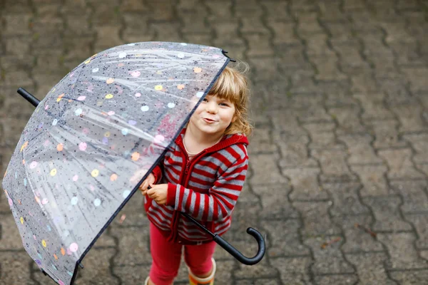 雨の日に大きな傘で遊んでいる小さな幼児の女の子。雨の中を走る幸せな正の子、水たまり。雨の服やゴムブーツを持つ就学前の子供。悪天候の日の子供の活動. — ストック写真