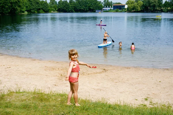 Malá blonďatá školačka se baví cákáním v létě v jezeře, venku. Šťastné dítě se učí plavat. Aktivní volný čas s dětmi na dovolené. Nebezpečí na domácích jezerech — Stock fotografie