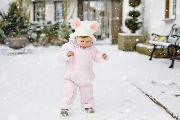 Gelukkig klein meisje maken eerste stappen buiten in de winter door de sneeuw. Leuke peuter die leert lopen. Kind heeft plezier op koude besneeuwde dag. Het dragen van warme baby roze kleding snowsuit en bobbels hoed. — Stockfoto