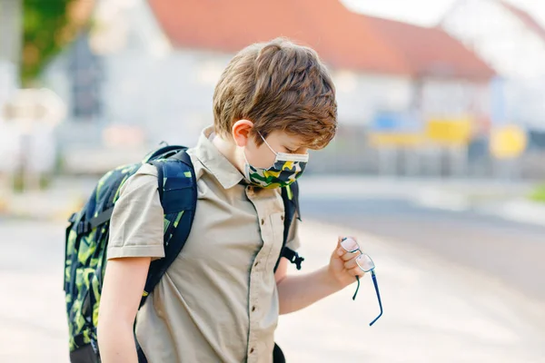 Corona virüsü salgını yüzünden gözlüklü ve tıbbi maskeli mutlu çocuk. Güneşli bir günde okul otobüsü bekleyen çantalı bir okul çocuğu. Sağlıklı bir çocuk sokakta.. — Stok fotoğraf