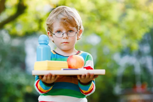 Förvirrad upprörd liten förskola pojke med böcker, äpple och dricka flaska på sin första dag till grundskolan. Sorgligt barn, student med glasögon, utomhus. Tillbaka till skolan, rädsla, utbildningskoncept. — Stockfoto