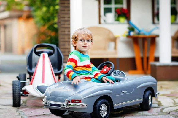 Menino pré-escolar dirigindo carro de brinquedo grande e se divertindo, ao ar livre. Criança feliz brincar e ter atividade ao ar livre. Criança com óculos sorrindo, ações de verão para crianças — Fotografia de Stock