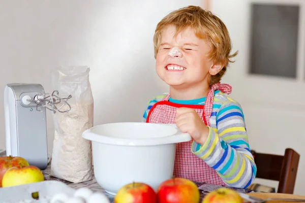 Bonito pequeno menino pré-escolar loiro feliz cozinhando bolo de maçã e muffins na cozinha doméstica. Engraçado adorável criança saudável se divertindo com o trabalho com misturador, farinha, ovos, frutas. Pequeno ajudante dentro de casa — Fotografia de Stock