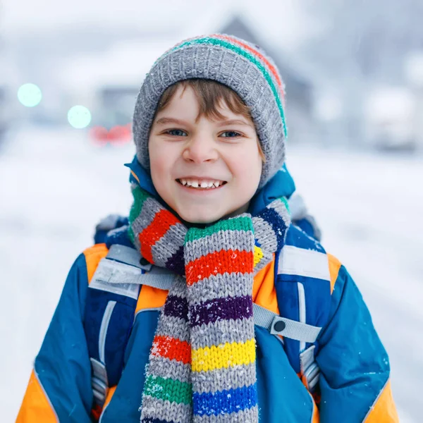 Menino da escola de classe primária a caminhar para a escola durante a queda de neve. Criança feliz se divertindo e brincando com a primeira neve. Estudante com mochila ou mochila em roupas coloridas de inverno. — Fotografia de Stock