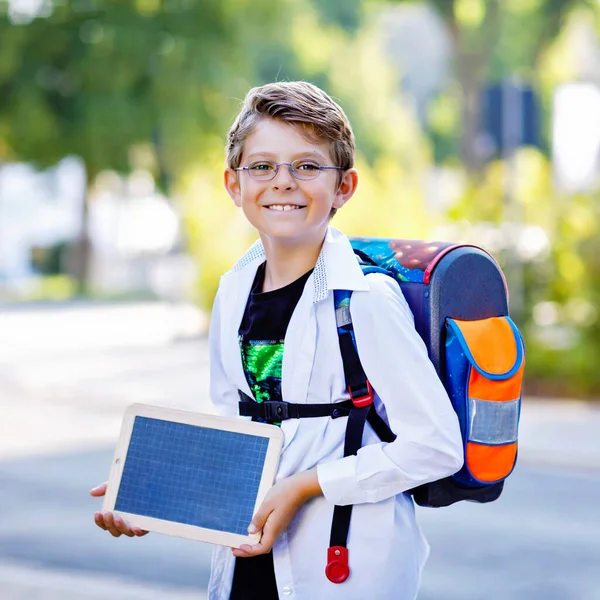Happy little kid boy with glasses and backpack or satchel. Schoolkid on the way to school. Healthy adorable child outdoors. Empty chalk desk in hands for copyspace and free text. Back to school.