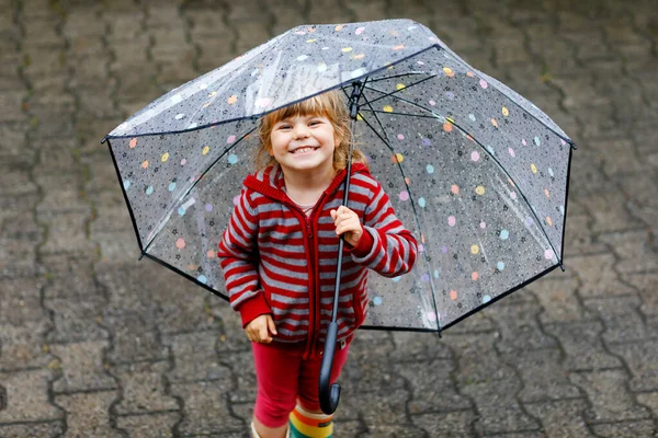 Little toddler girl playing with big umbrella on rainy day. Happy positive child running through rain, puddles. Preschool kid with rain clothes and rubber boots. Children activity on bad weather day. — Stock Photo, Image
